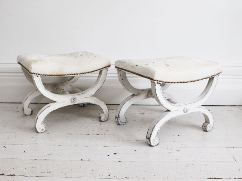 A pair of 19th C white painted scroll legged stools upholstered in cowhide